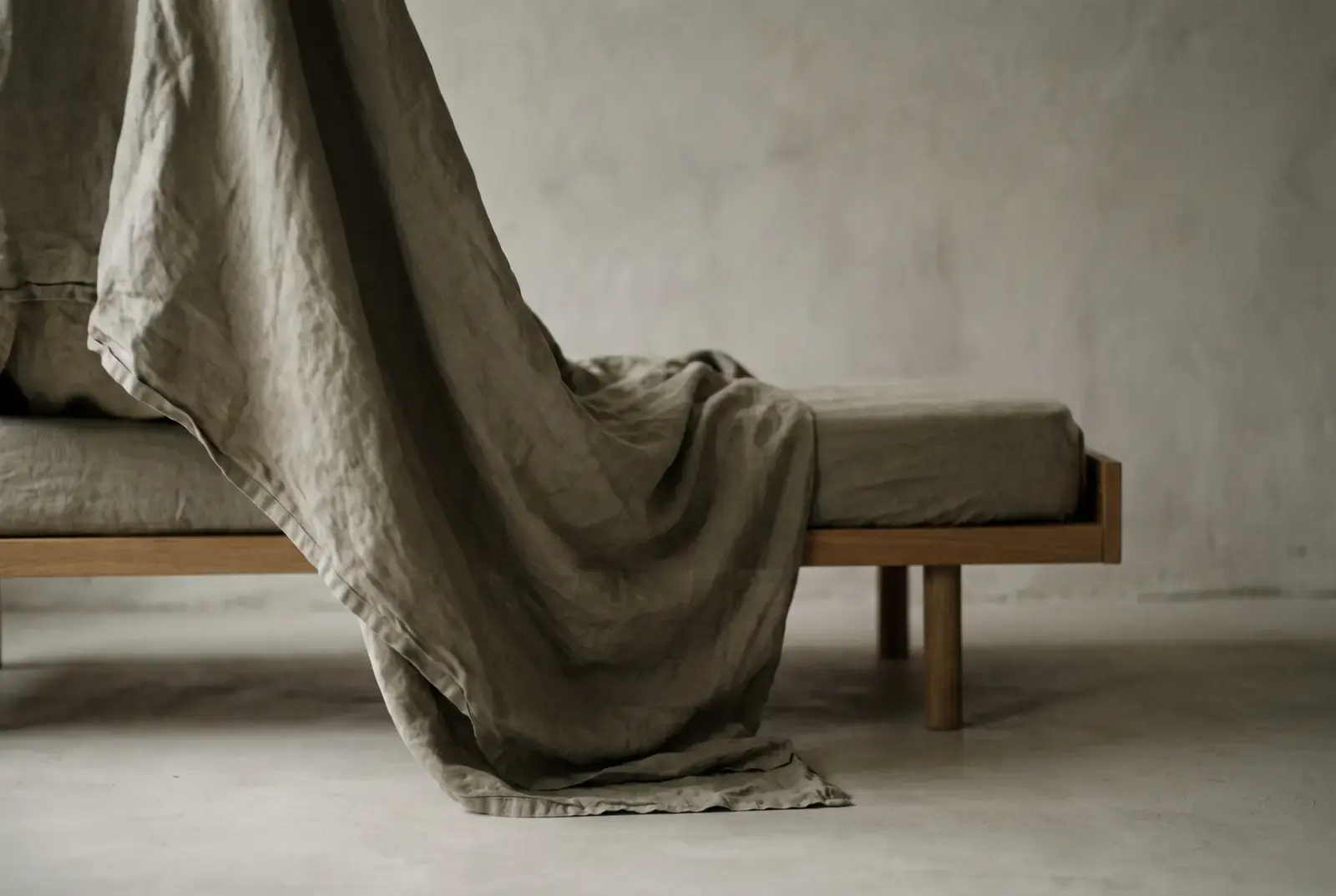 A heavy linen sheet trailing from an unmade daybed onto a pale floor in low morning light