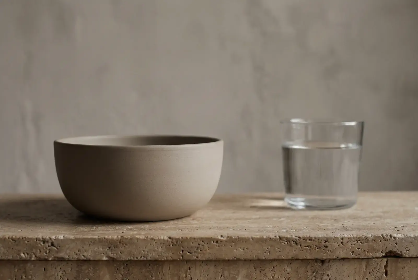 A plain unglazed ceramic bowl and a glass of water on a travertine ledge in soft daylight