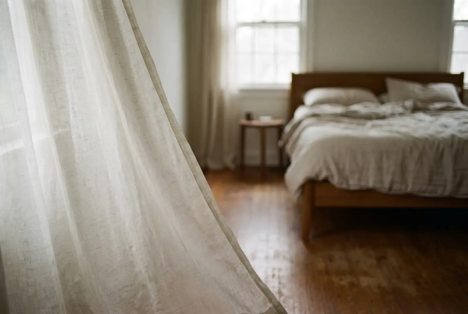 Early daylight through a sheer linen curtain in a quiet bedroom, the bed still unmade