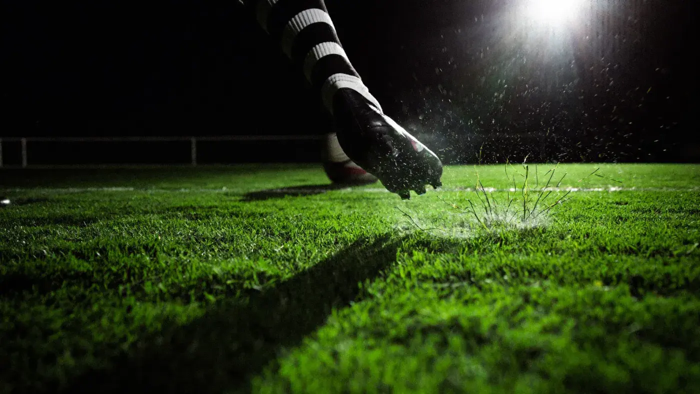 A boot striking a ball on floodlit turf at night