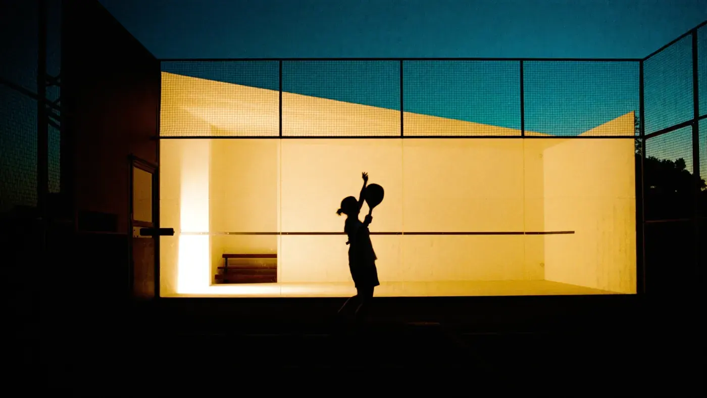 A padel player in silhouette against a floodlit glass court at dusk